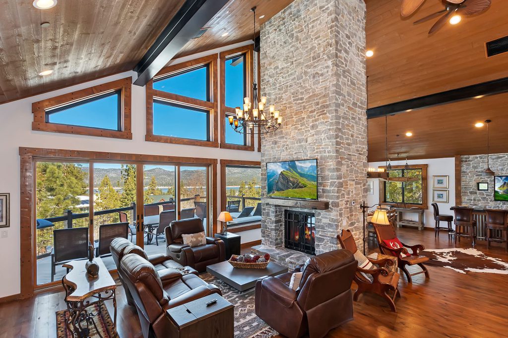 A luxurious and spacious living room in a Big Bear cabin with a two-story stone fireplace, large windows overlooking the lake, and multiple seating areas.