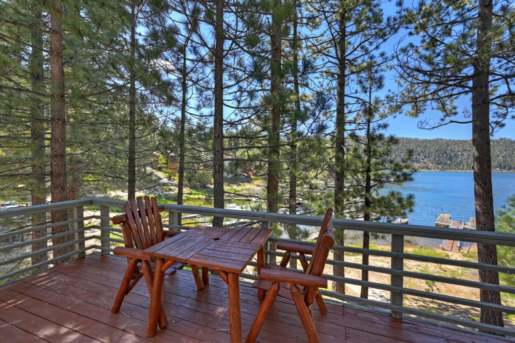 Wooden cabin deck with chairs and table overlooking a peaceful lake surrounded by pine trees.