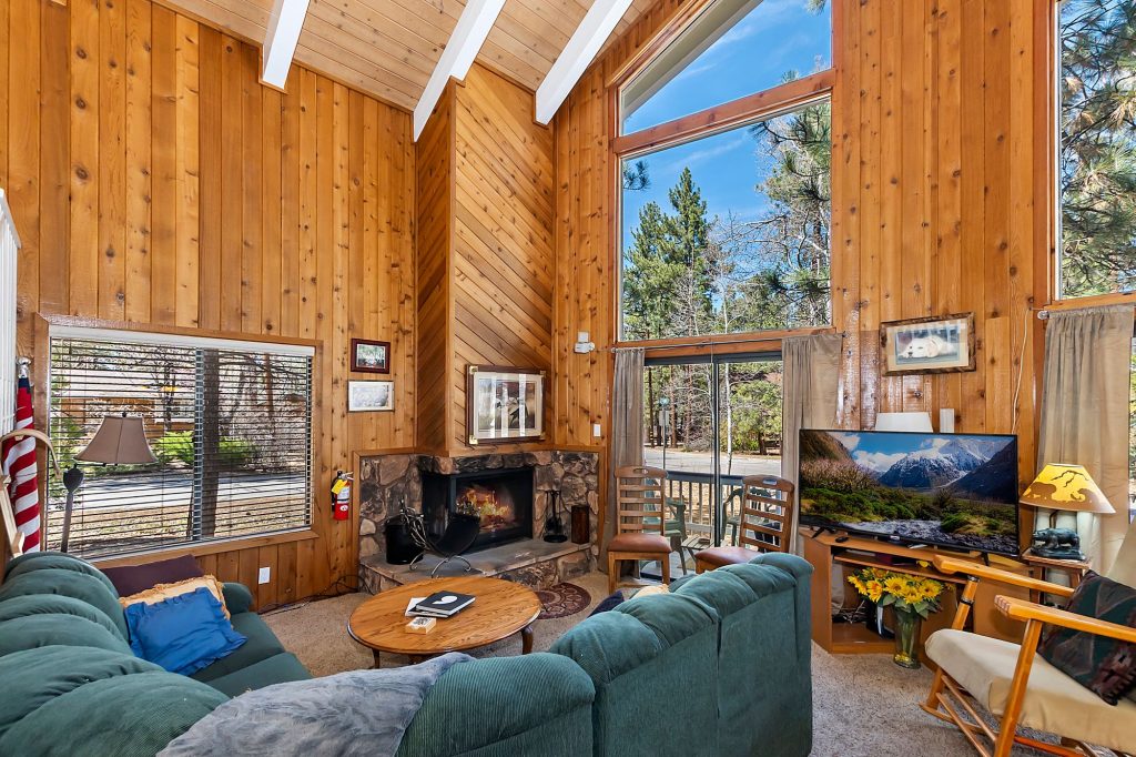 Whispering Cottage living room with wooden walls, stone fireplace, large windows, cozy seating, and forest views.
