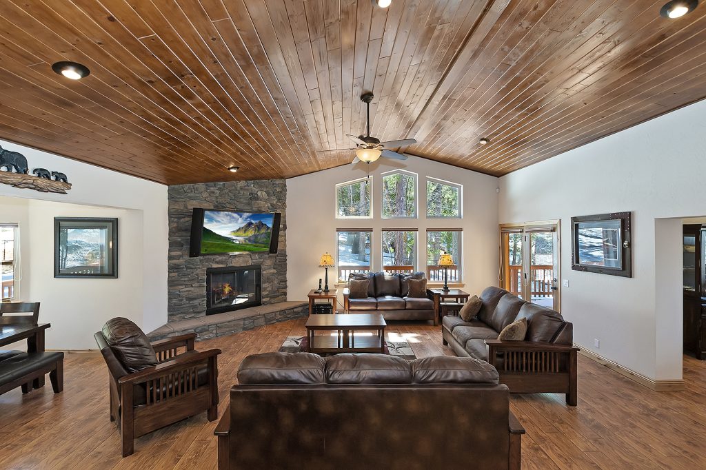 Spacious cabin living room with stone fireplace, wood ceiling, leather seating, and forest views