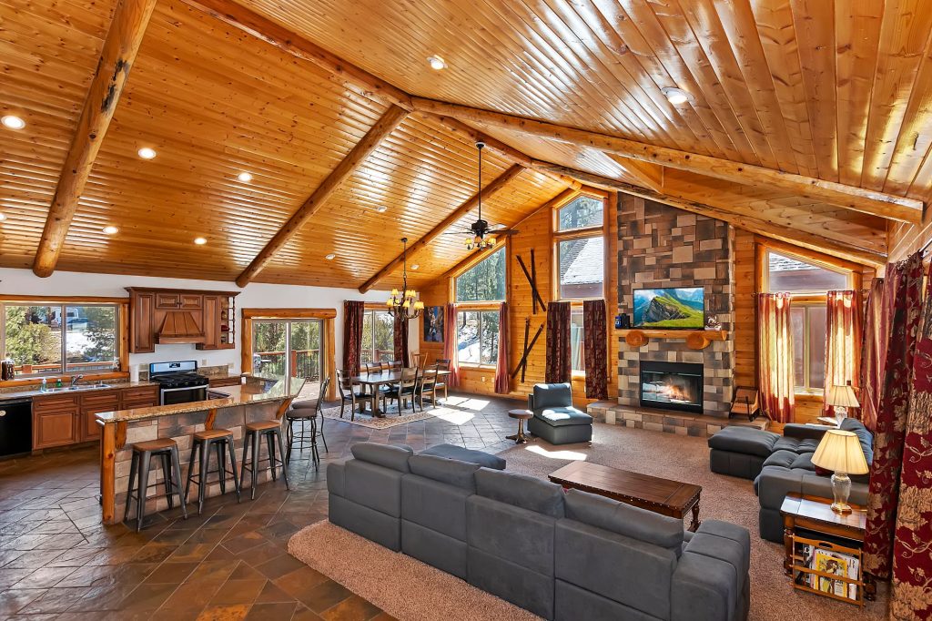 Spacious Serenity Now Lodge interior with a wood-beamed ceiling, stone fireplace, kitchen, dining area, and comfortable seating
