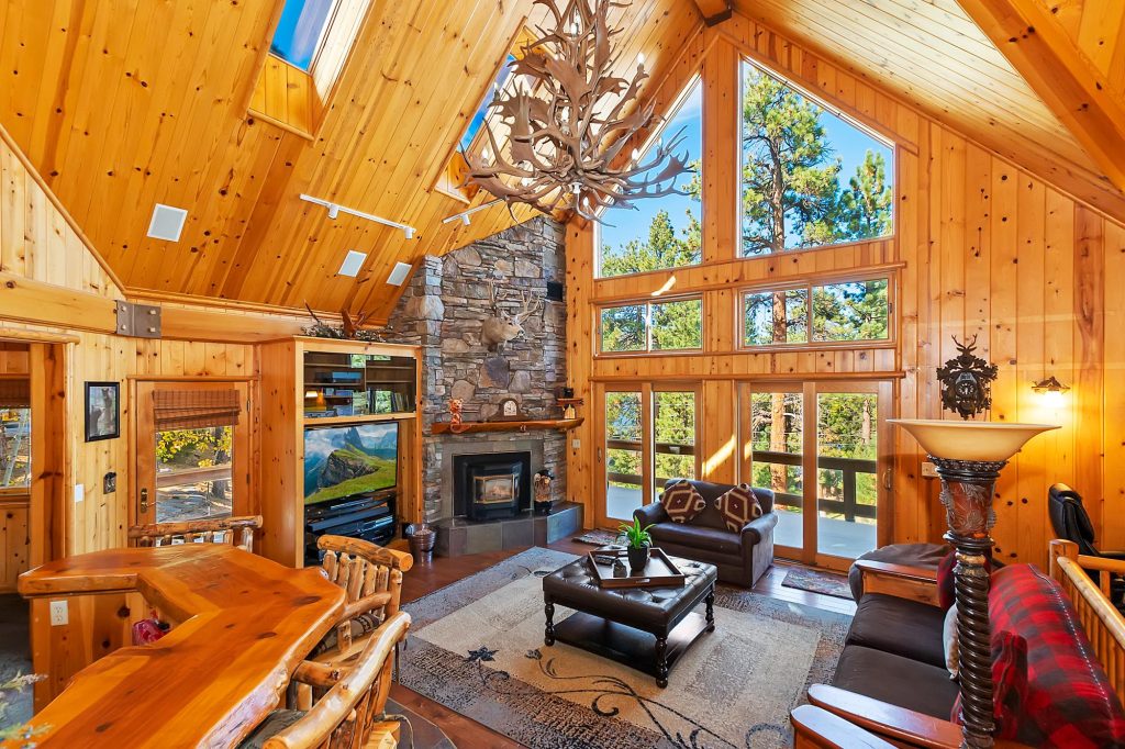 Cozy cabin living room at Waterview Retreat featuring wood interiors, stone fireplace, large windows, and forest views in Big Bear Lake