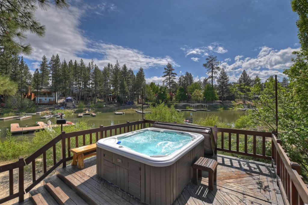 Outdoor hot tub on a wooden deck overlooking a lake with docks and surrounding pine trees under a partly cloudy sky.