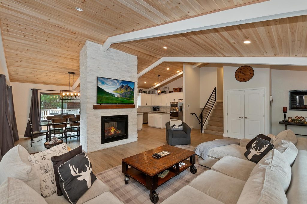 Modern cabin living room at Dew Drop Inn featuring a stone fireplace, wood ceiling, cozy seating, and open kitchen area in Big Bear Lake, CA
