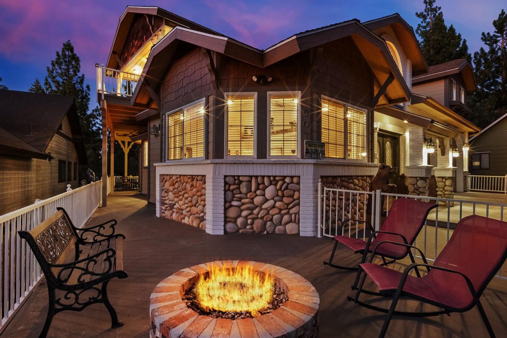 Spacious bedroom with vaulted ceilings, cozy armchairs, four-poster bed, and red wood stove fireplace, opening to a balcony with forest views.