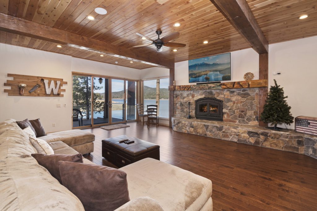 A living room with a large stone fireplace, a brown sectional sofa, and a wood ceiling has a large glass door that opens to a patio and a lake in Big Bear.