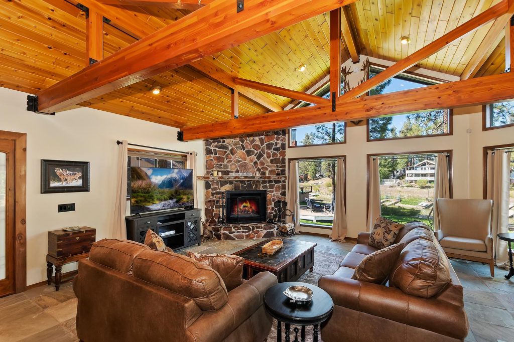 Interior living room with vaulted wood ceiling, stone fireplace, and large windows overlooking the lake.