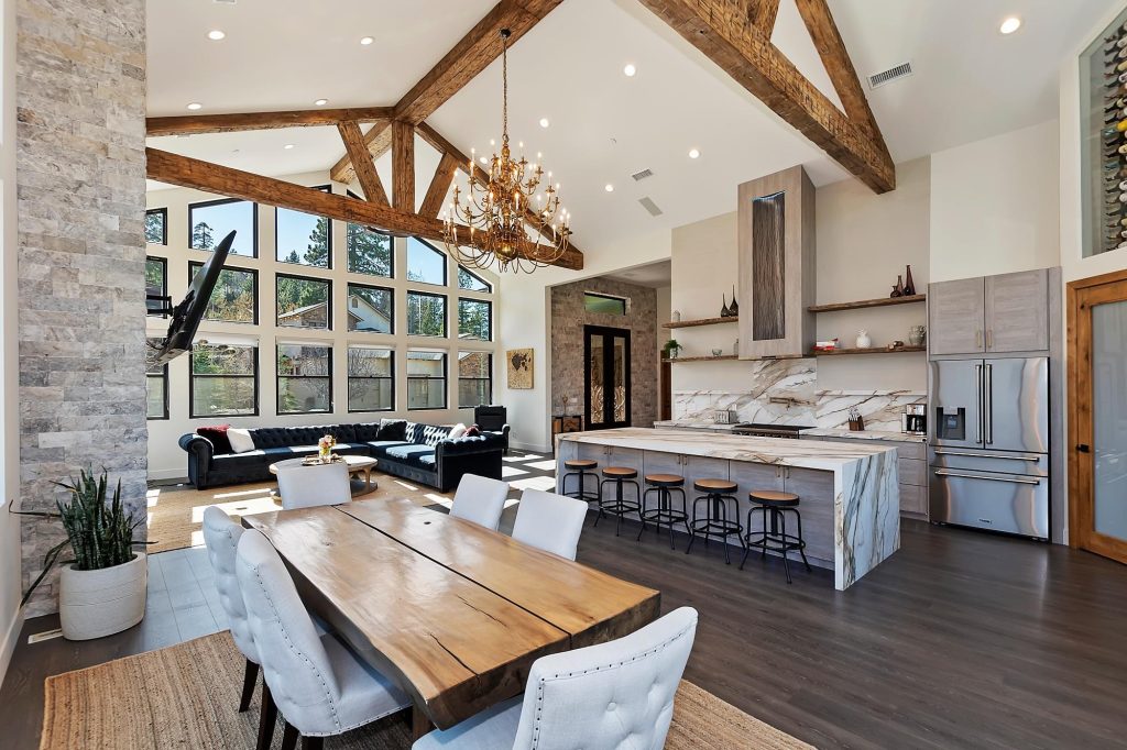 Spacious modern open-concept living room and kitchen with vaulted ceilings, rustic wooden beams, large windows, and a stylish chandelier