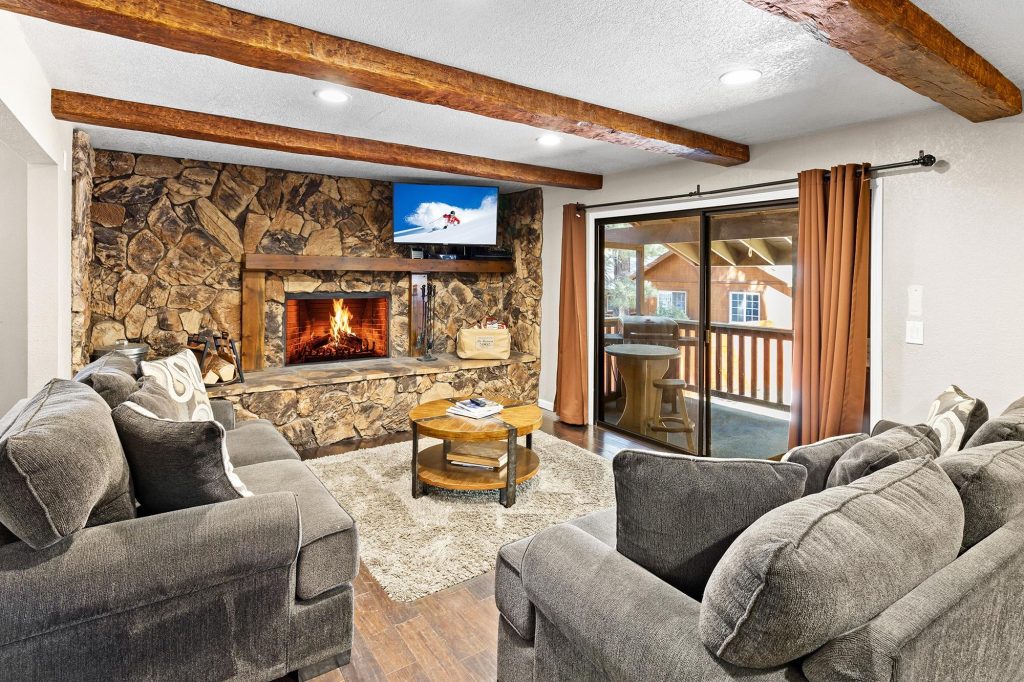 Cozy living room with gray sofas, stone fireplace, wooden beams on ceiling, and mounted TV above fireplace