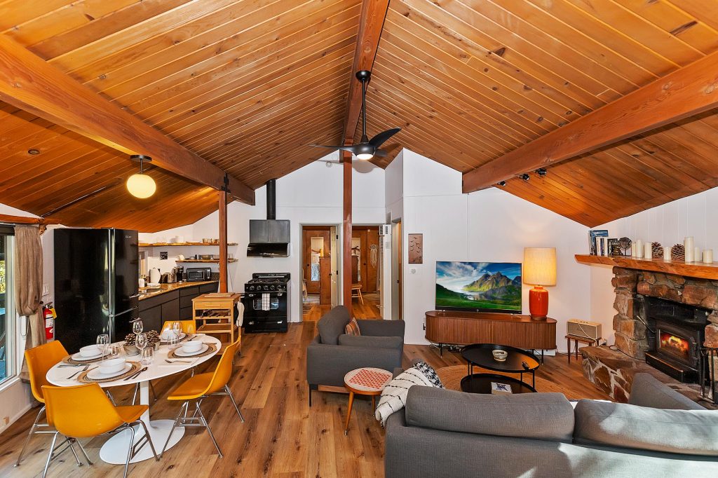 Rustic cabin interior with wood ceiling, fireplace, kitchen area, and cozy living space