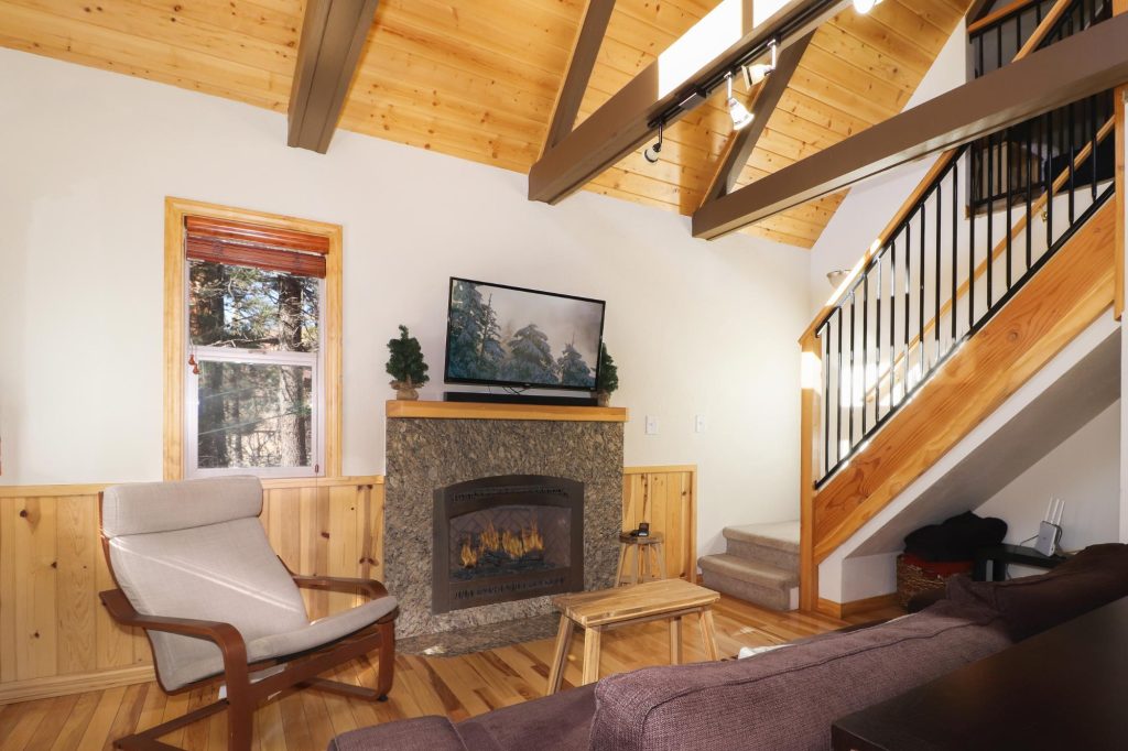 Cozy cabin living room with stone fireplace, wood ceiling, staircase, and comfortable seating