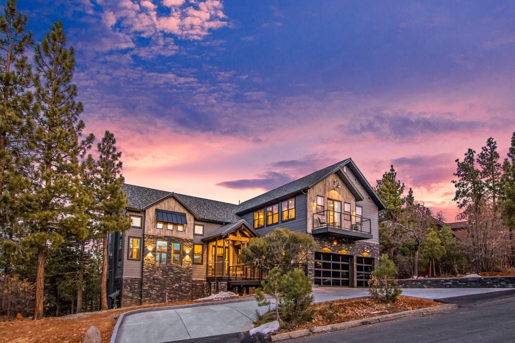 A modern cabin-style house with a multi-car garage, surrounded by pine trees under a vibrant sunset sky in Big Bear.