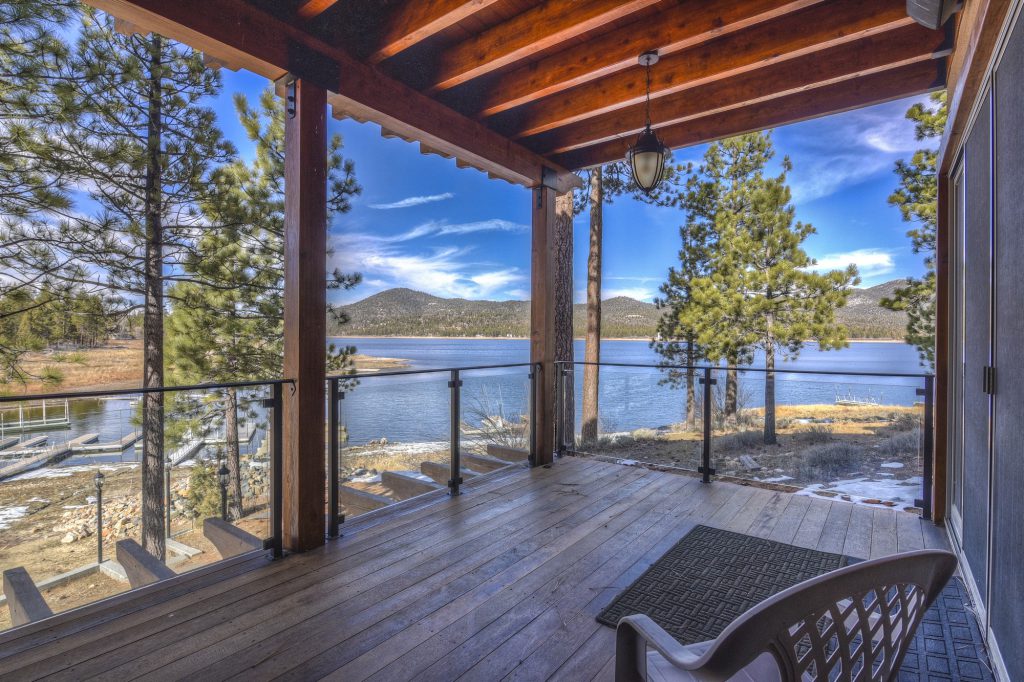 A covered wooden deck with a glass railing offers a winter view of Big Bear Lake and the surrounding mountains.