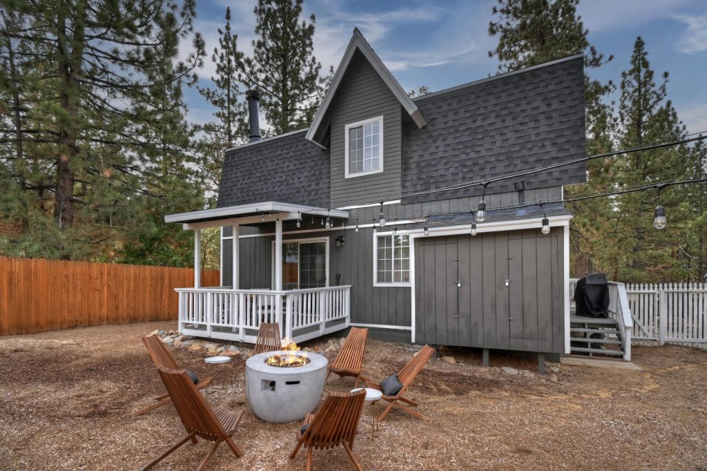 Modern dark-gray cabin in Big Bear surrounded by pine trees, featuring a backyard fire pit with wooden chairs and outdoor string lights.