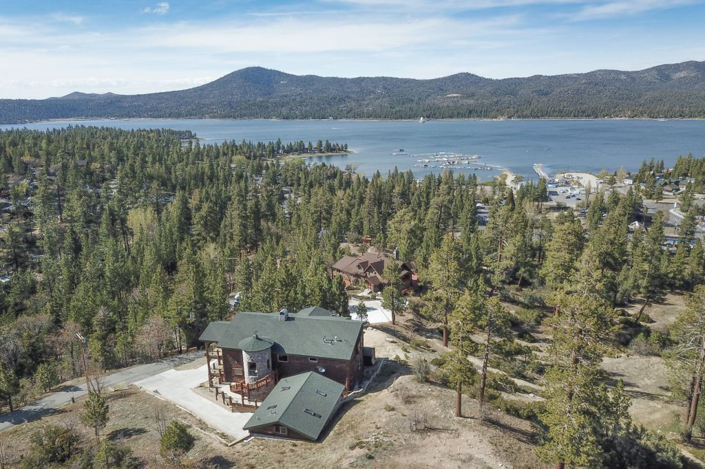 An aerial view shows a house nestled in a pine forest with a clear view of Big Bear Lake and mountains.