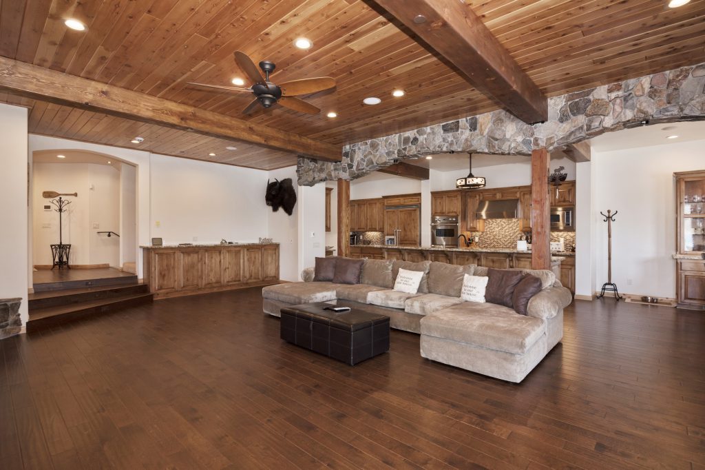A large, open-plan living area with a brown sectional couch, a dark wood floor, and a wood and stone ceiling is visible, with a kitchen in the background in Big Bear.