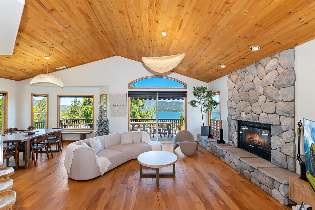 Interior living room with wood ceiling, stone fireplace, and large windows framing Big Bear Lake view.