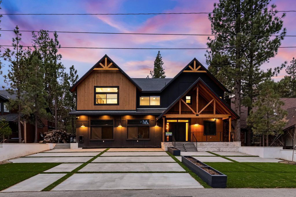 A beautiful, modern, two-story cabin with a concrete driveway and a manicured lawn is nestled among pine trees at sunset in Big Bear.