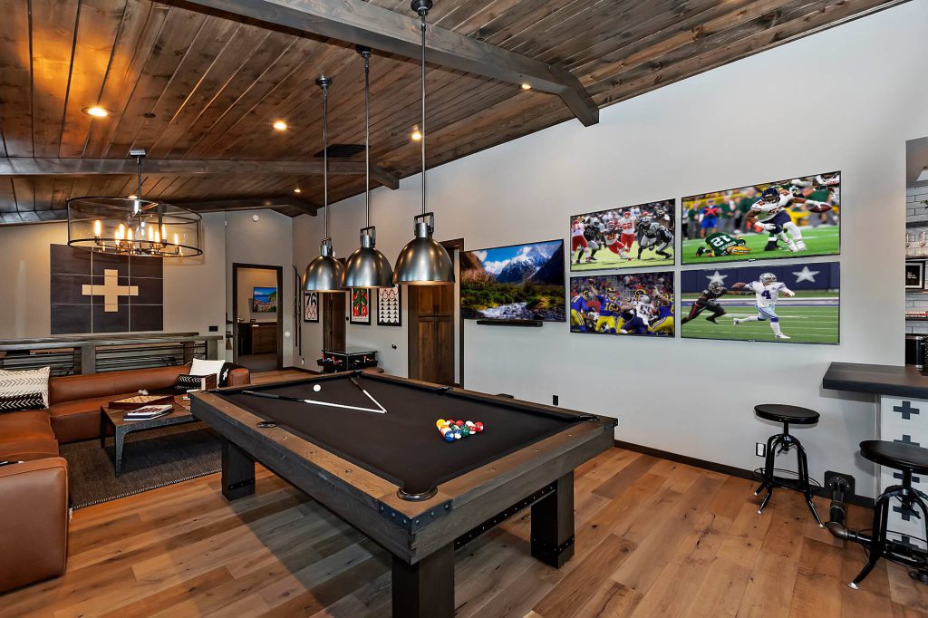 The interior of a Big Bear cabin's game room with a pool table, multiple TVs showing a football game, and rustic decor.