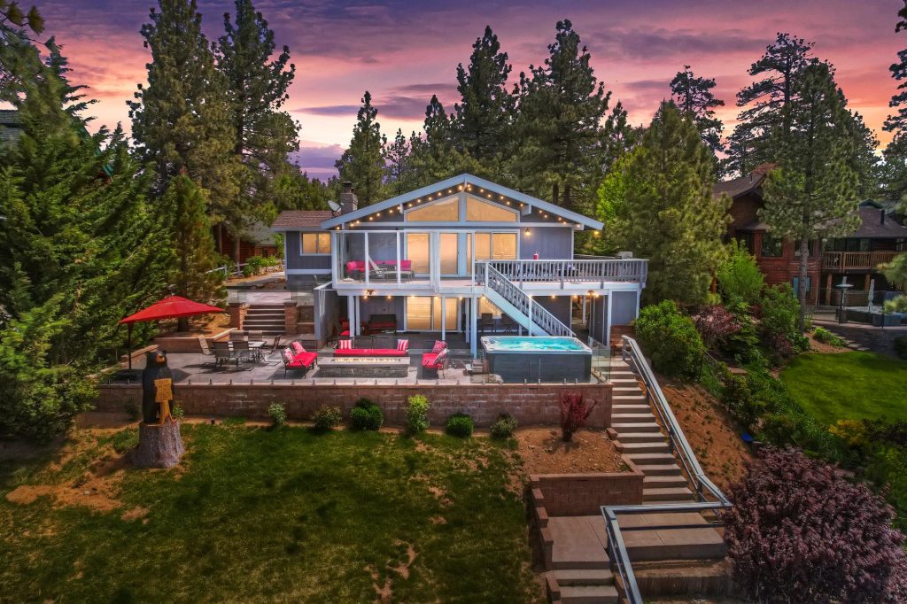 Two-story house exterior at twilight with deck, hot tub, patio furniture, and surrounding pine trees.