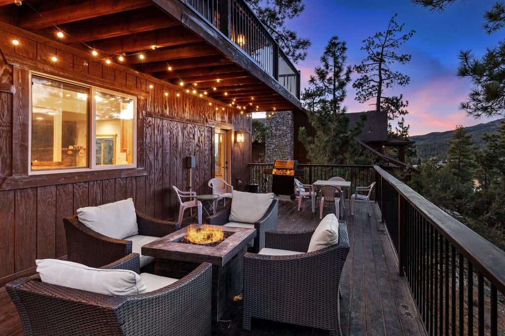 Evening view of Mountain Chic cabin in Big Bear featuring a spacious deck with string lights, outdoor fire pit seating, and forest views.