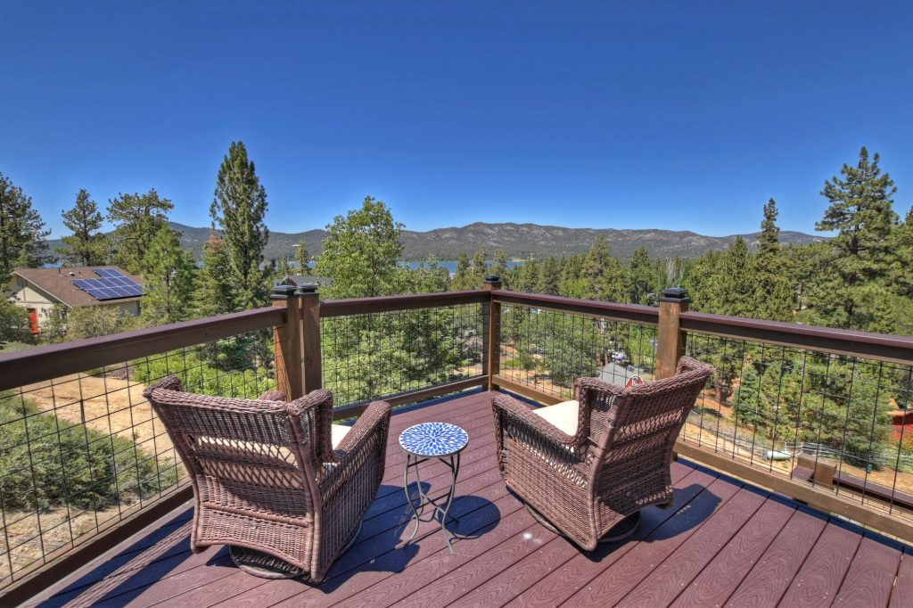 Outdoor deck with two wicker chairs, small round table, and a railing overlooking a forest and distant mountains under a clear blue sky.