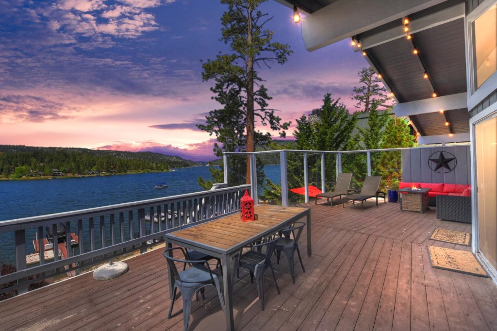 Spacious balcony overlooking a serene lake with patio furniture, trees, and a colorful sunset sky.