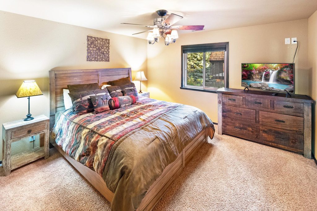 Cozy bedroom with queen bed, ceiling fan, lamps, and a window showing greenery outside.