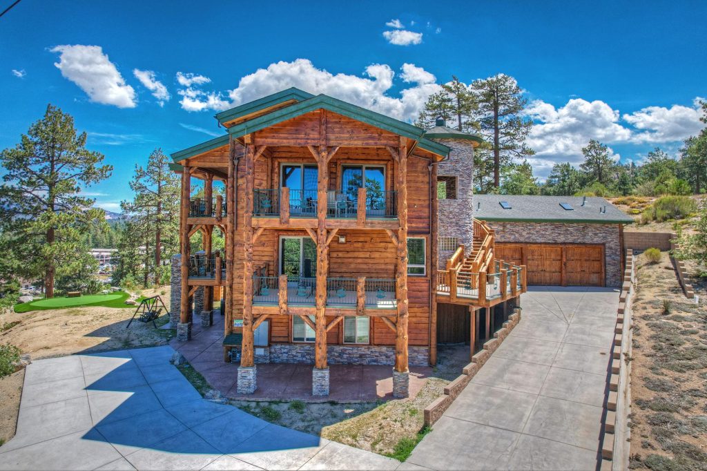 Exterior view of a multi-story log cabin with stone accents, balconies, and surrounding pine trees under a partly cloudy sky.