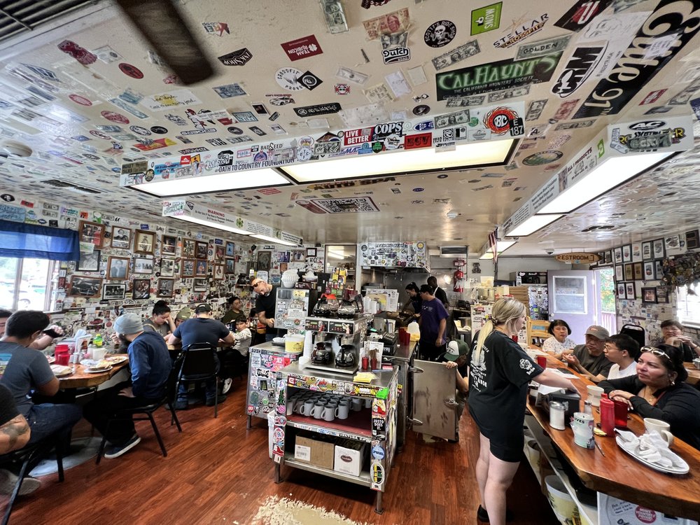 An interior shot of a lively, rustic diner with walls and ceilings covered in stickers, license plates, and photos, and people dining at tables and a counter in Big Bear.