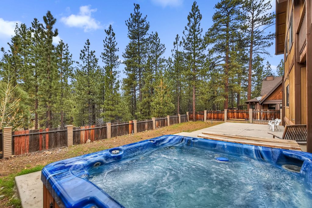 Backyard scene with a hot tub in the foreground, tall pine trees, and a fenced yard on a clear sunny day