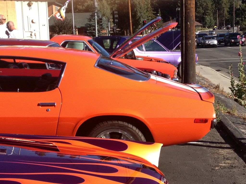 A row of classic cars is parked at the Big Bear car show, including an orange car with white racing stripes and a purple one with flames.