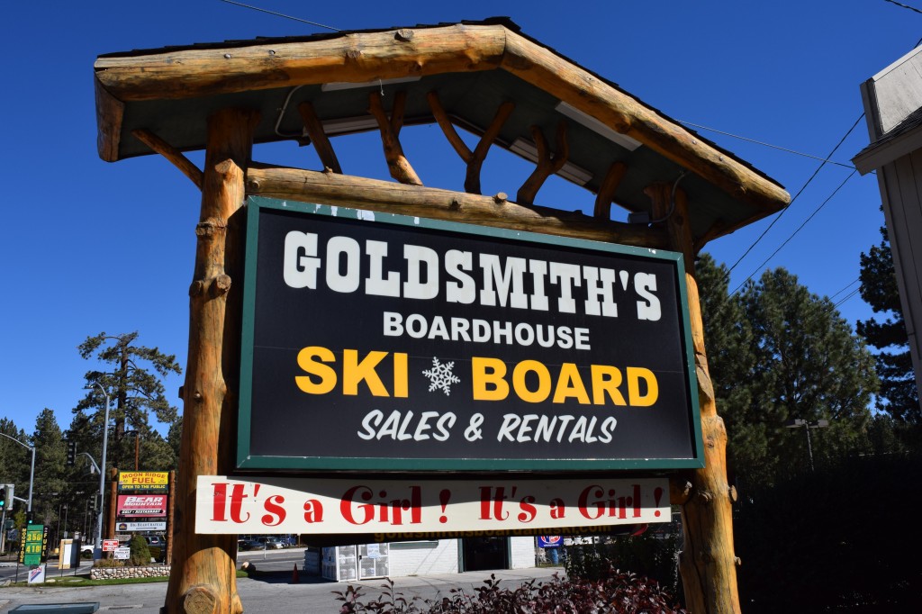An outdoor wooden sign for "Goldsmith's Boardhouse Ski & Board Sales & Rentals" in Big Bear, with a small sign below it that says, "It's a Girl!"