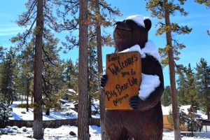 A large snow-dusted bear statue holding a "Welcome to Big Bear Lake" sign stands among pine trees in Big Bear.