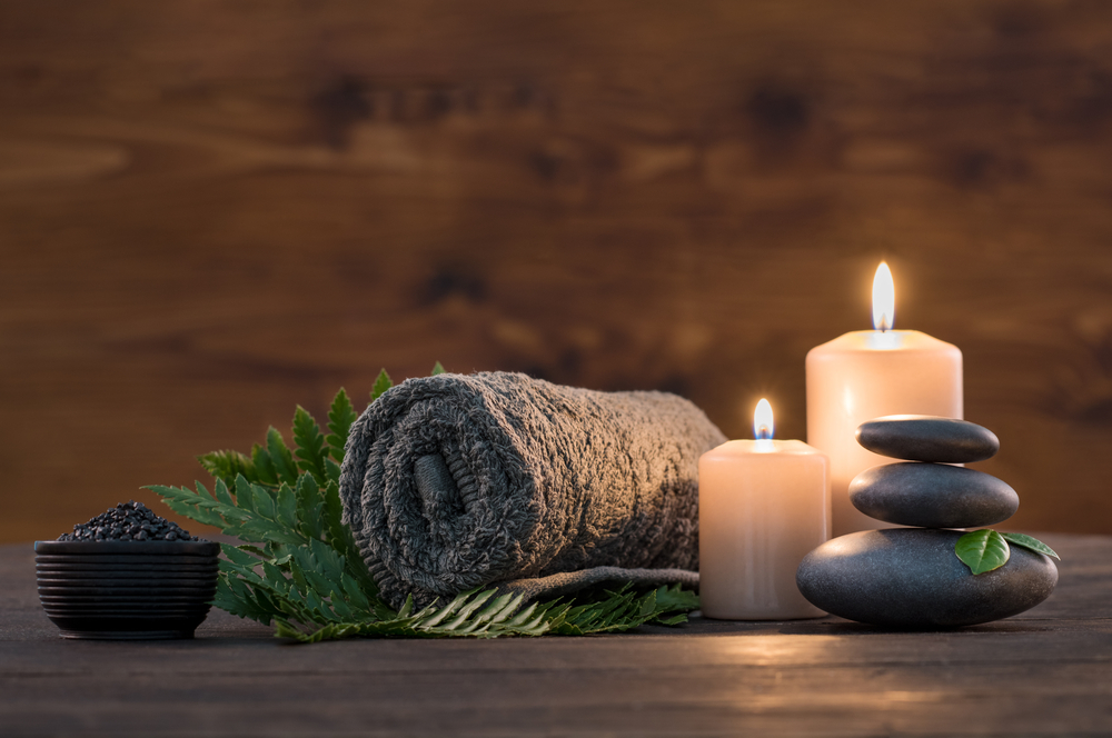 A tranquil spa setup with a rolled towel, burning candles, a stack of smooth stones, and a bowl of bath salts against a wooden background.