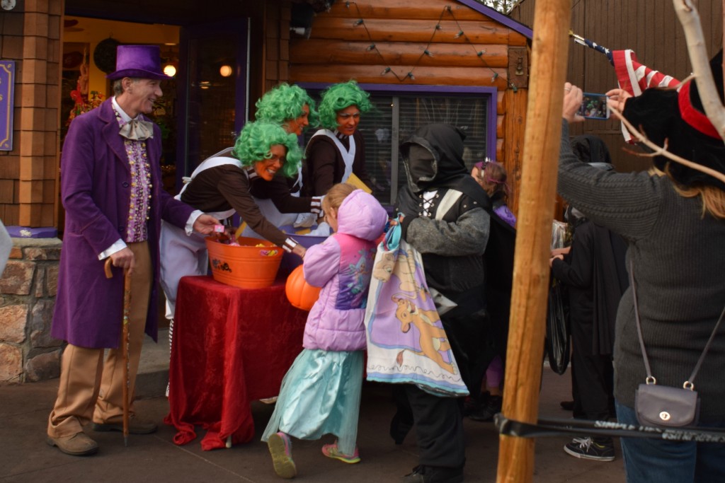 A festive Halloween scene at Big Baer, featuring a smiling Willy Wonka, Oompa Loompas, and children receiving treats.