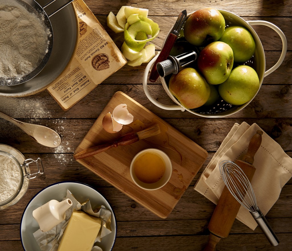 A top-down view of baking ingredients on a rustic wooden table, including flour, eggs, butter, apples, and various kitchen tools like a rolling pin and whisk.