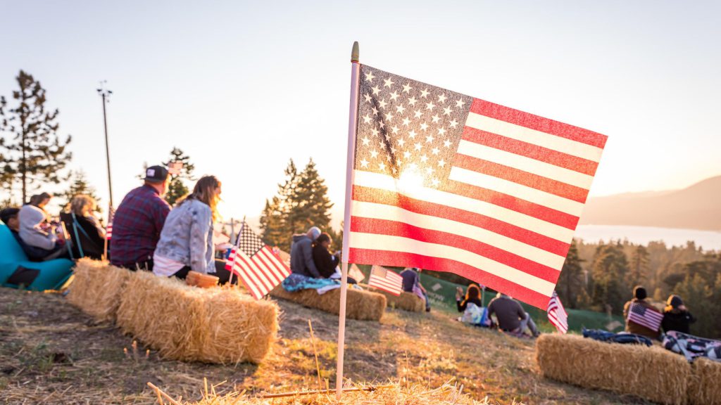 People sit on hay bales on a grassy hill with an American flag, watching the sunset at Big Bear.