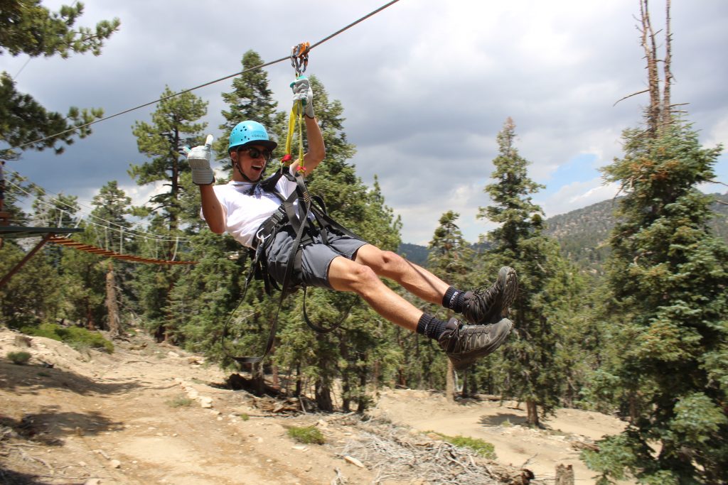A person smiling and giving a thumbs up while ziplining through a forest at Big Bear.