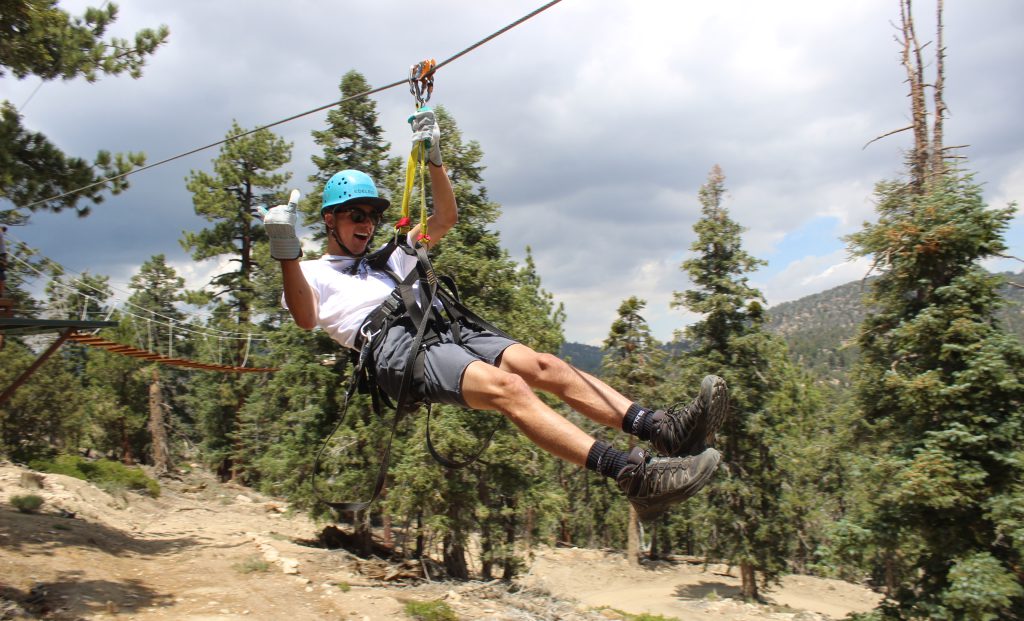 A person in a blue helmet and harness smiles while ziplining through a forest with pine trees under a cloudy sky.