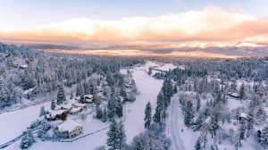 A high-angle view of a snow-covered forest and cabins at Big Bear, with a frozen lake running through the center and a stunning sky with pink and orange clouds overhead at Big Bear