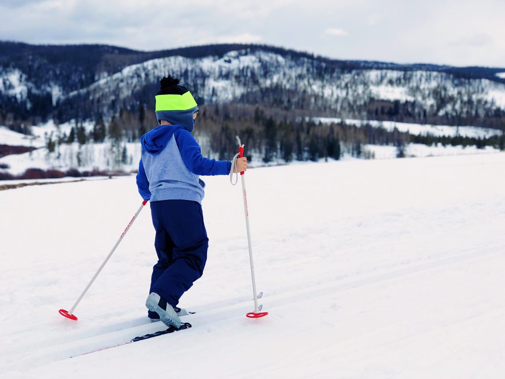 A child wearing a blue and gray jacket and a beanie is cross-country skiing on a snowy field with a mountain in the background at Big Bear.