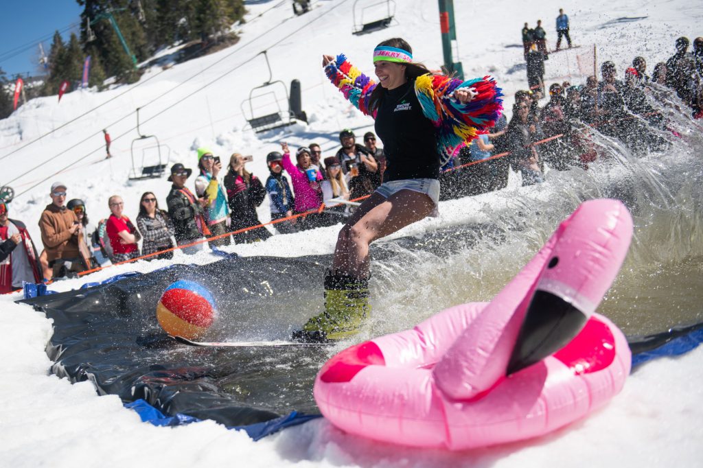 Snowboarder splashing through a pond skim pool during Bear Break at Big Bear Mountain Resort while spectators cheer.