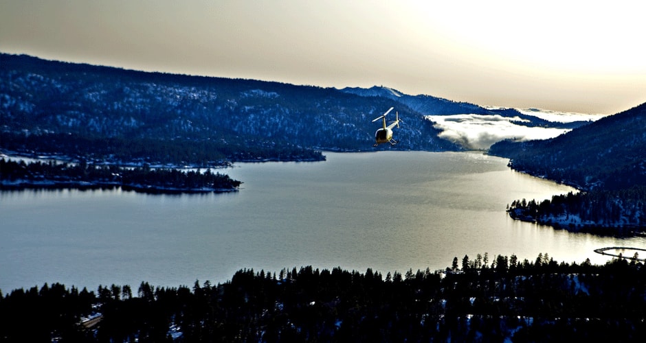 A ski lift hovers over Big Bear Lake, surrounded by snow-dusted mountains and evergreen trees during a scenic helicopter tour.
