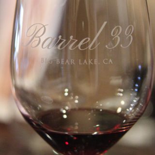 A close-up shot of a wine glass with a small amount of red wine, showing the "Barrel 33, Big Bear Lake, CA" logo etched on the glass.