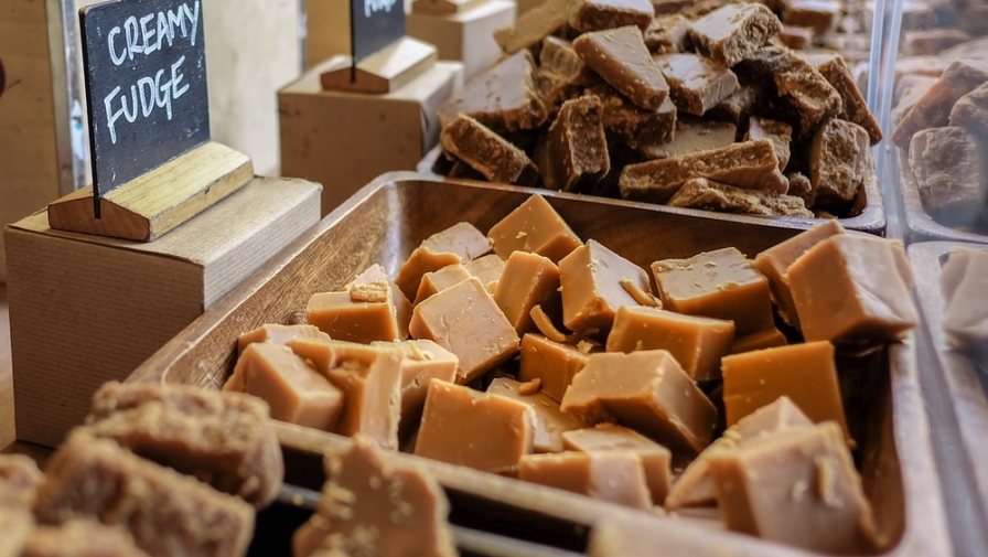 A close-up of a large wooden tray filled with chunks of creamy fudge, with a sign in the background that reads "CREAMY FUDGE".