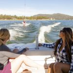 Two women on a speedboat at Big Bear Lake watch a friend wakeboard, surrounded by scenic pine-covered hills.