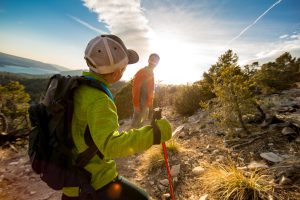 Two hikers ascend a sunny, rocky trail at Big Bear with a lake and mountains in the distant background.