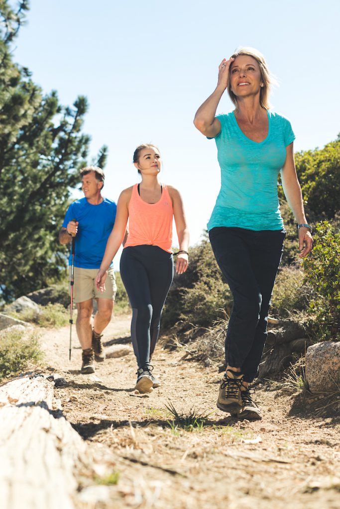 Three people are hiking on a sunny trail at Big Bear, with one woman adjusting her hat.
