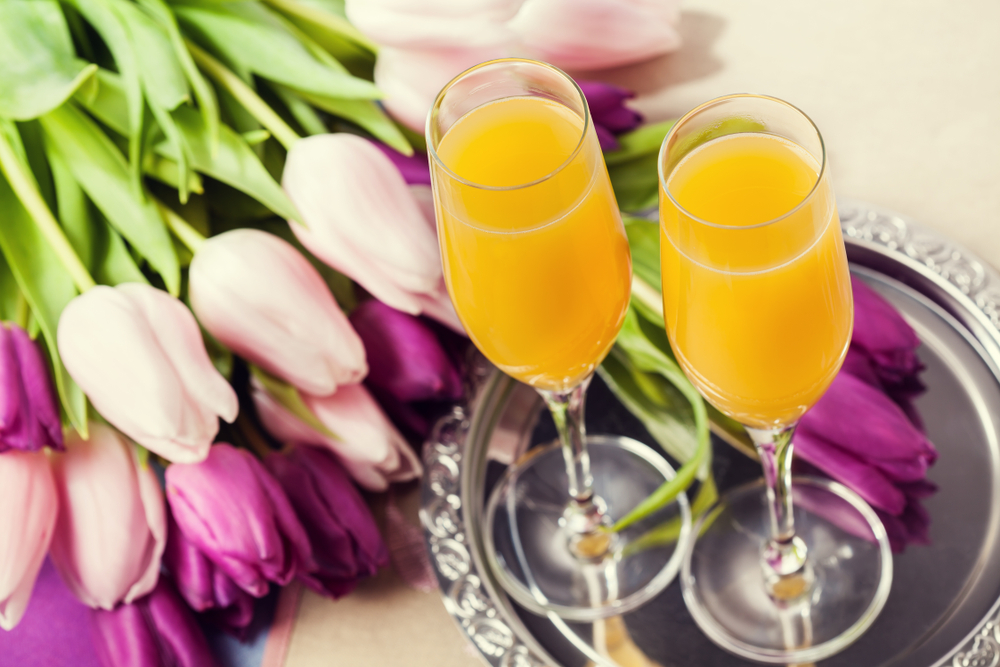 Two flutes of orange mimosas on a silver tray next to a bouquet of pink and purple tulips.